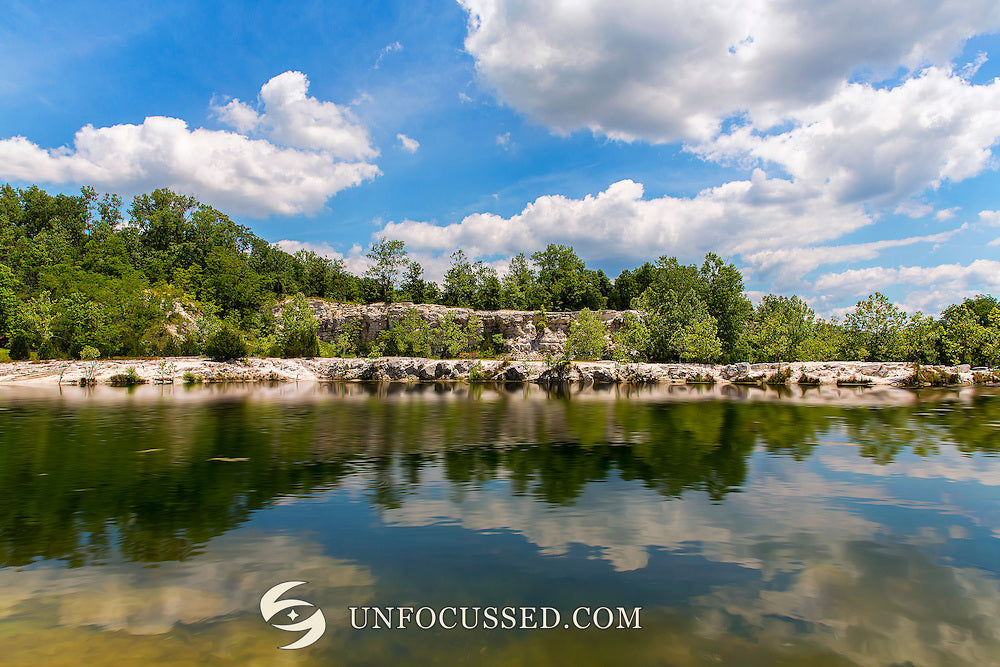 Alberi verdi e una riva rocciosa chiara riflessi nel lago di Klondike Park sotto cielo blu e nuvole soffici.