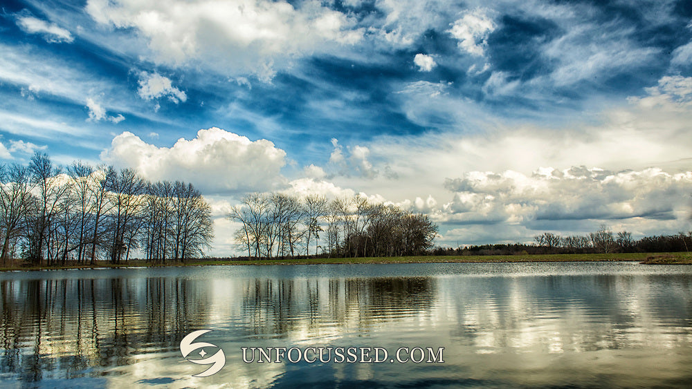 Alberi spogli e nuvole bianche riflessi in un ampio lago sotto un cielo grigio-blu materico.