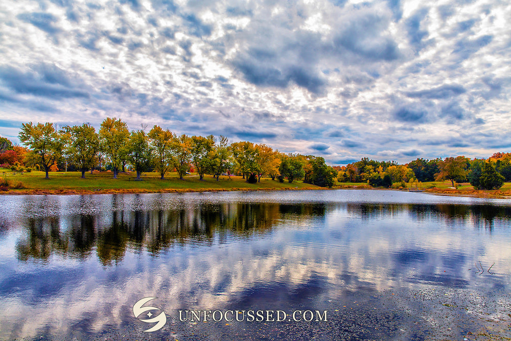 Textured white and gray clouds reflect in a blue lake bordered by green and autumn-colored trees.