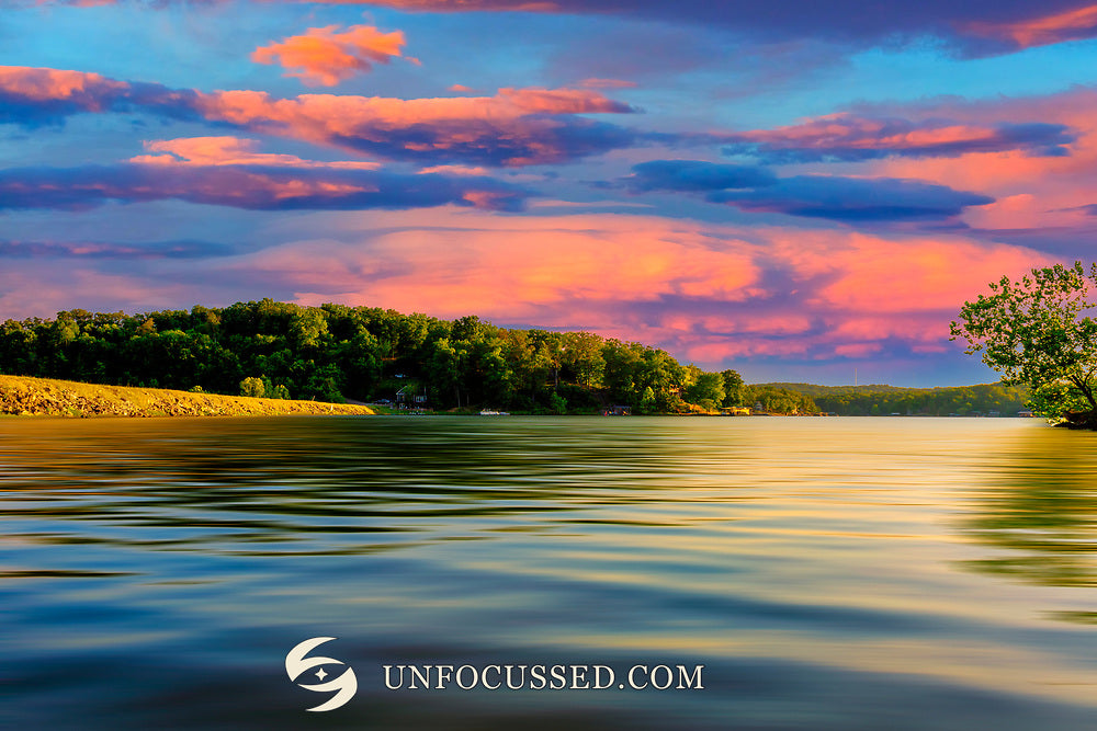 Pink and coral clouds above Goose Creek Lake and its dam, with blue rippled water, a golden shoreline and green trees.