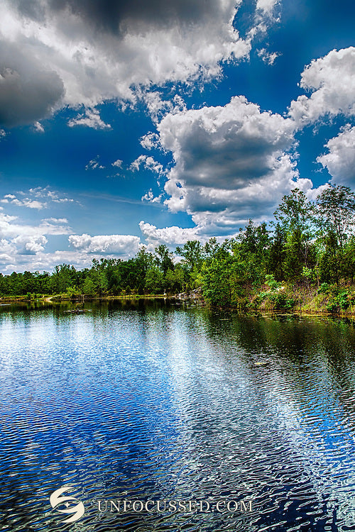 Une eau bleu profond ondulée sous des arbres verts et de grands nuages blancs, dans une vue verticale du lac de Klondike Park.