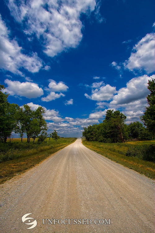 A pale gravel road narrowing between green trees and grasses beneath a tall blue sky scattered with white clouds.