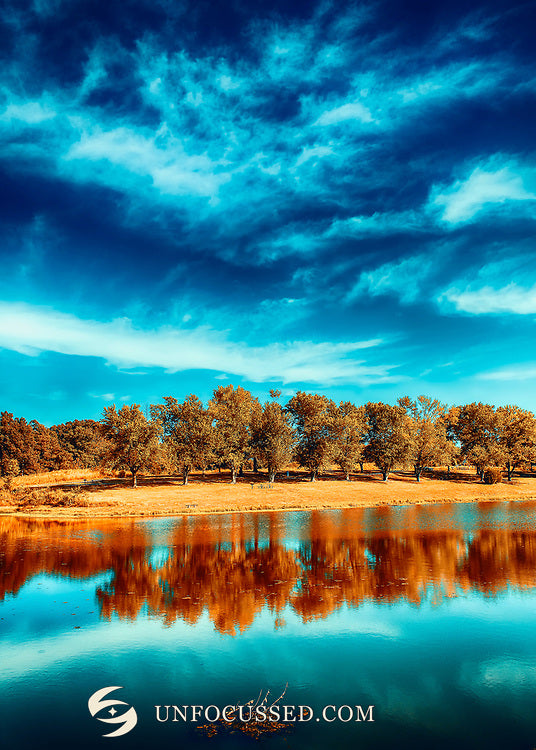 Goldene Herbstbäume und ihre Spiegelung unter dramatischem türkisfarbenem Himmel in einer Seeansicht im Hochformat.