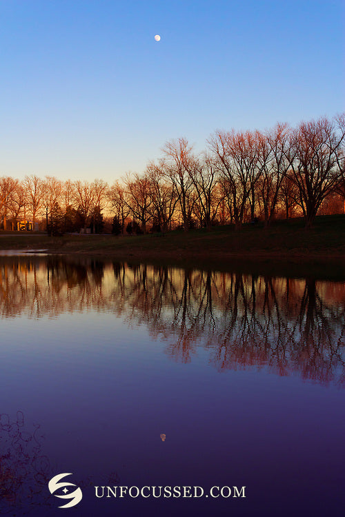 A small moon hangs above bare lakeside trees, with warm horizon light and purple-blue water reflections.